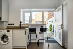 A compact kitchen with a washing machine, two bar stools, and a window overlooking a courtyard. - Immaculate 6-bed House in Newcastle (Wallsend)