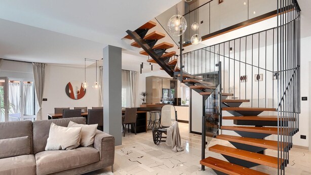 A modern living room with a grey sofa, a dining area, and a staircase with wooden steps and metal railings. - Stunning home in Pula (Pula)