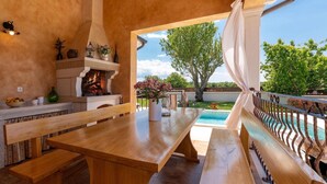 An outdoor patio with a wooden table and benches, a fireplace, and a view of a swimming pool and trees. - Amazing home in Porec (Porec)