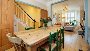 A dining area with a wooden table and chairs, a staircase, and a living room with a sofa and a lamp. - Sunshine Joy, by Veeve (London)