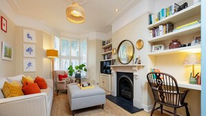A living room with a sofa, ottoman, and a fireplace. There are bookshelves and a chair. - Sunshine Joy, by Veeve (London)