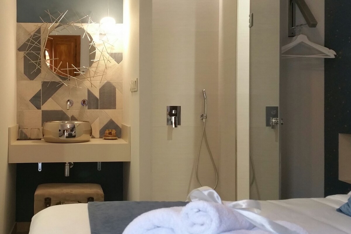 A hotel room with a bed, a bathroom with a shower, a sink with toiletries, and a mirror with a geometric design.