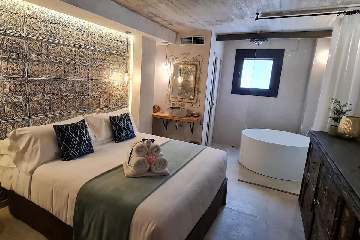 Un dormitorio moderno con pared estampada, una cama con almohadas, una mesita de noche, un espejo, una ventana y un baño con lavabo e inodoro.