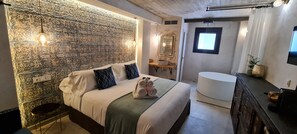 A modern bedroom with a patterned wall, a bed with pillows, a bedside table, a mirror, a window, and a bathroom with a sink and toilet. - Murallas de Jayran Hotel Boutique (Almeria)
