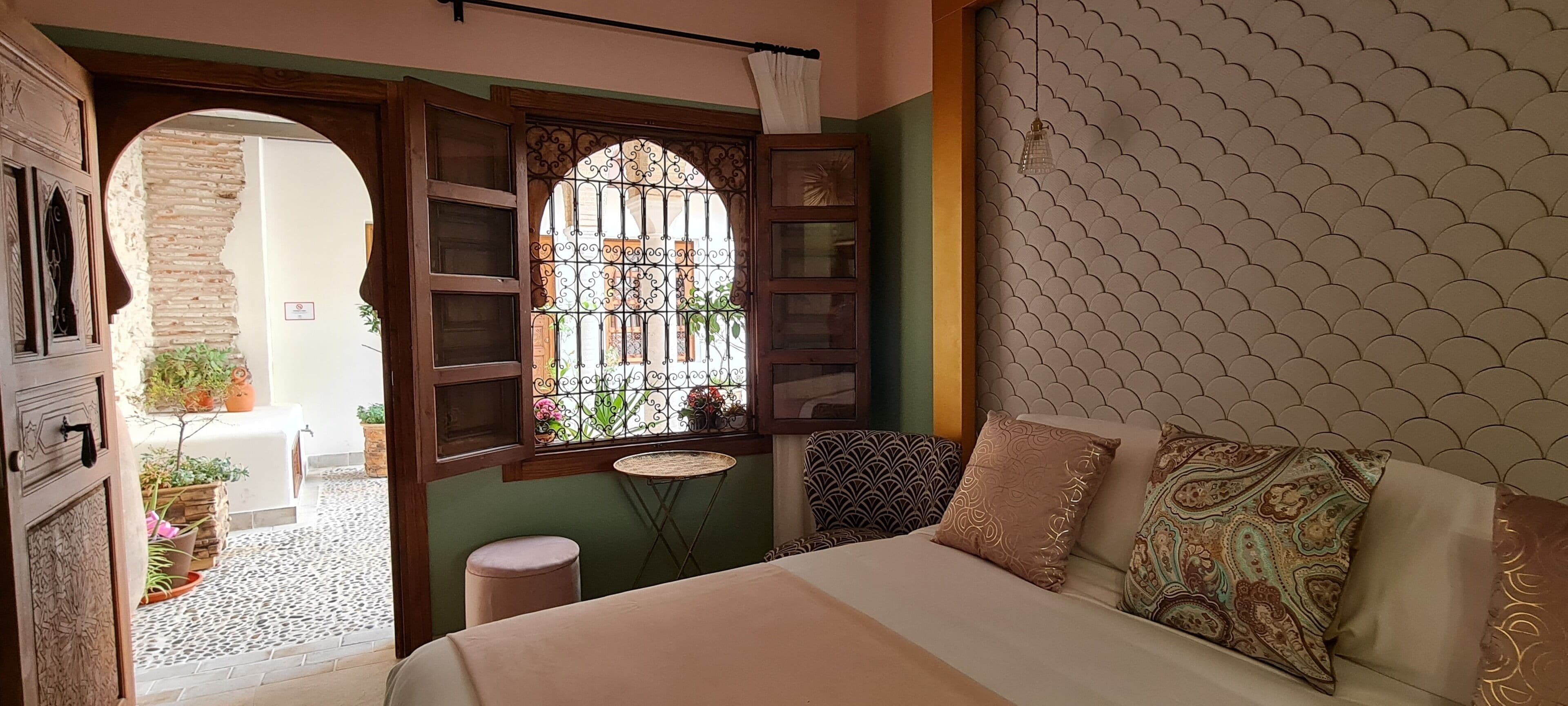 Ein Schlafzimmer mit einem Bett, einem Fenster mit schmiedeeisernen Gitterstäben, einem kleinen runden Tisch und einer Zimmerpflanze.