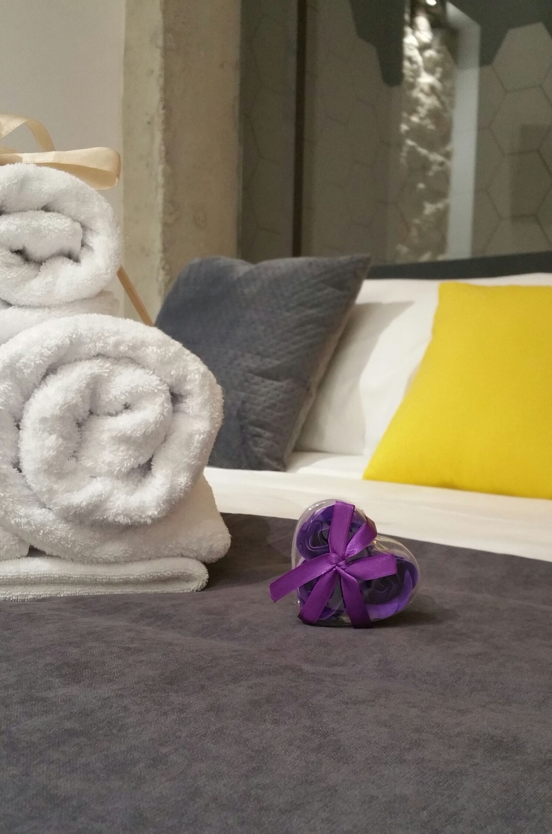A neatly made bed with a white and yellow pillow, a gray pillow, and a rolled white towel on a gray carpet.