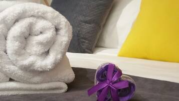 A neatly made bed with a white and yellow pillow, a gray pillow, and a rolled white towel on a gray carpet.