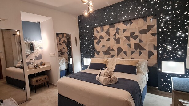 Un dormitorio moderno con cabecera de diseño geométrico, una mesita de noche con espejo y un pequeño rincón de descanso.