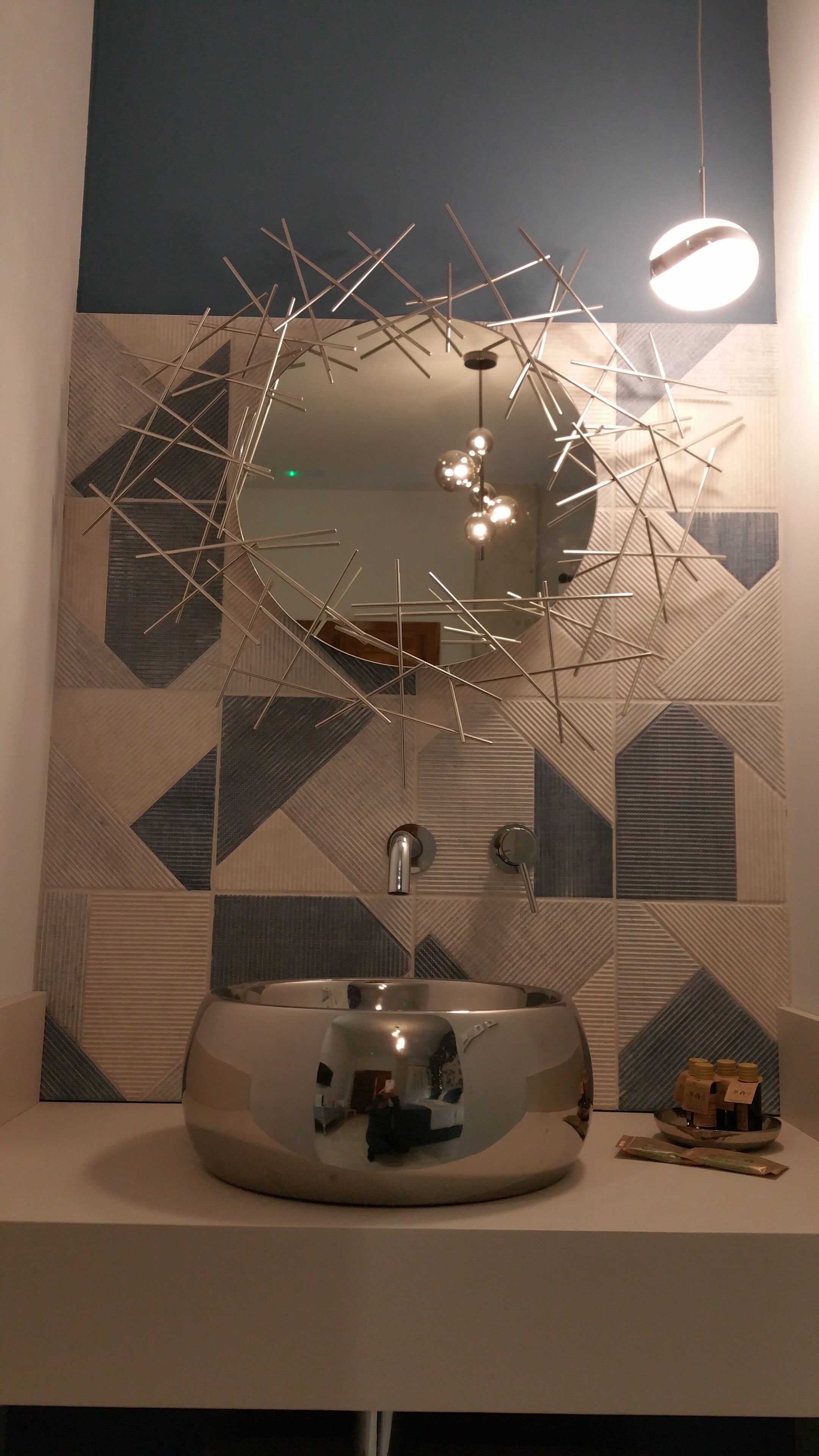 A modern bathroom with a geometric patterned wall, a round mirror, and a contemporary light fixture.