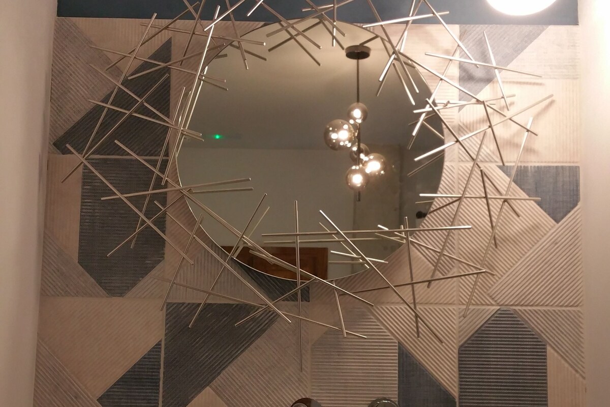 A modern bathroom with a geometric patterned wall, a round mirror, and a contemporary light fixture.