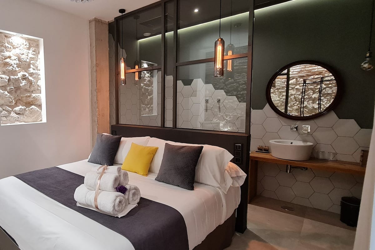 Una habitación de hotel moderna con una cama grande, un televisor montado en la pared, un ventanal con textura similar a la piedra y un baño con un espejo redondo y azulejos hexagonales.