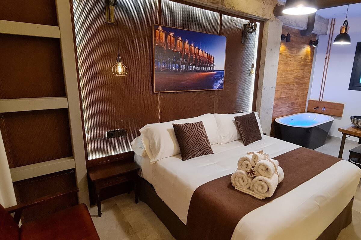 Habitación de hotel moderna con una cama grande, una mesita pequeña, una bañera y vistas a una construcción de madera.