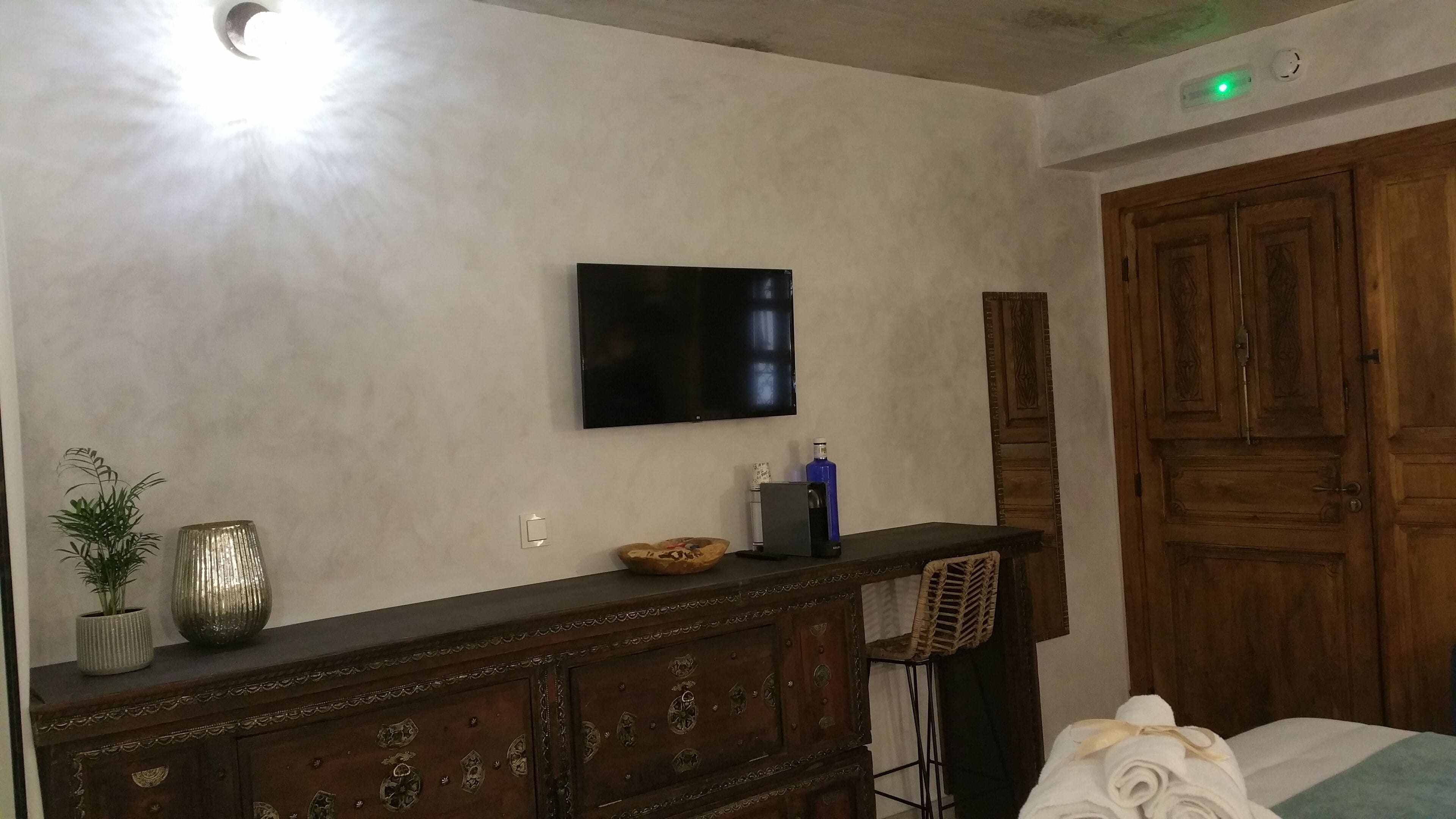 Ein Zimmer mit an der Wand montiertem Fernseher, einem Holzschrank mit verzierten Griffen, einer kleinen Pflanze und einer blauen Flasche auf der Ablagefläche.