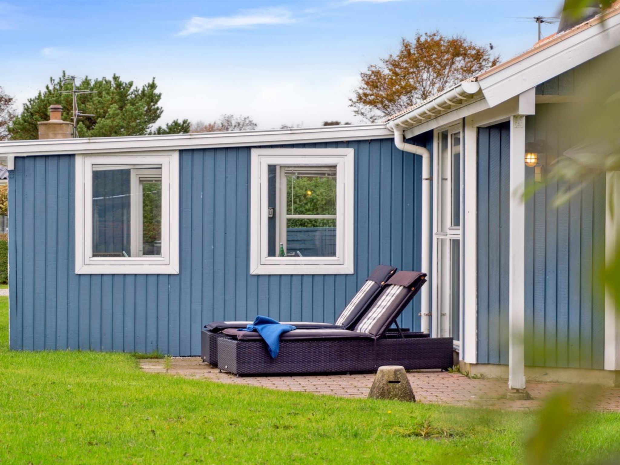 Ett litet blått hus med två fönster och en uteplats med en solstol och ett bord.