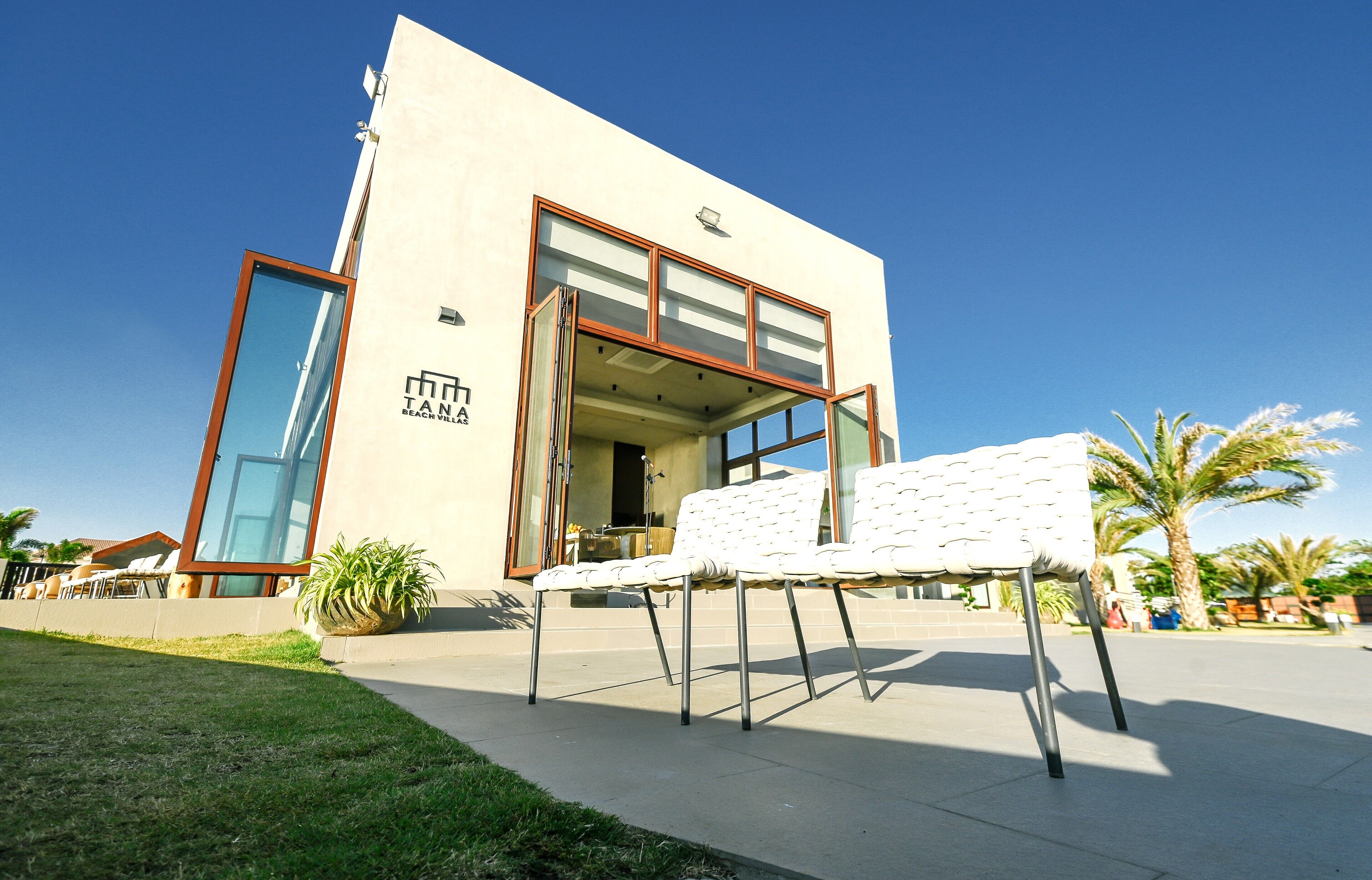 A modern building with a large glass entrance, a white outdoor seating area, and palm trees in the background.