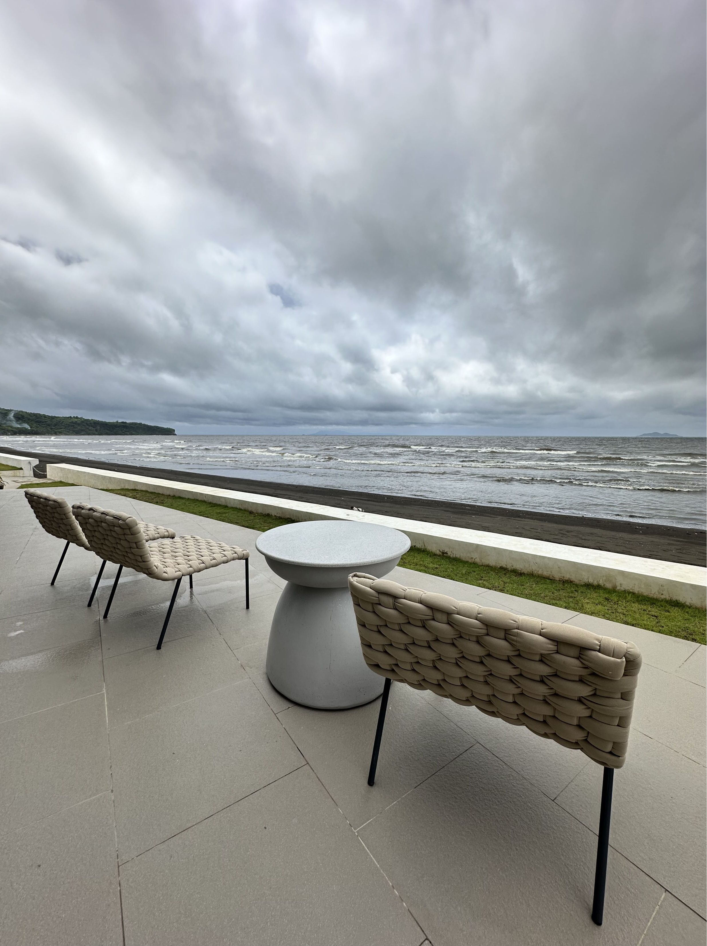Eine Terrasse am Meer mit zwei gepolsterten Stühlen und einem runden Tisch, mit Blick auf den Ozean unter einem bewölkten Himmel.