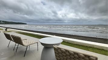 Eine Terrasse am Meer mit zwei gepolsterten Stühlen und einem runden Tisch, mit Blick auf den Ozean unter einem bewölkten Himmel.