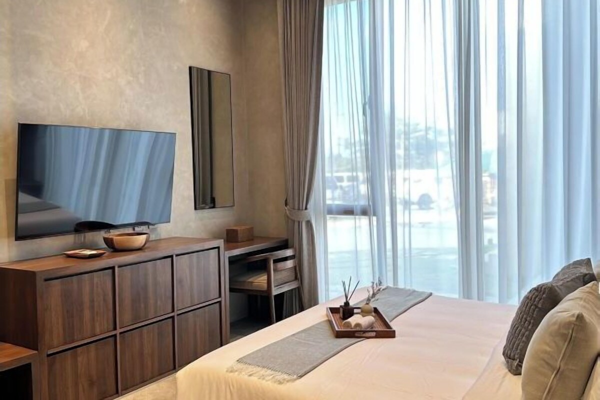 A modern hotel room with a large bed, a flat-screen TV, a wooden dresser, and a view of the city through large windows.