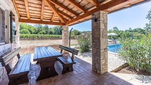 Un patio al aire libre con una mesa y bancos de madera, pilares de piedra, una piscina y un viñedo al fondo.