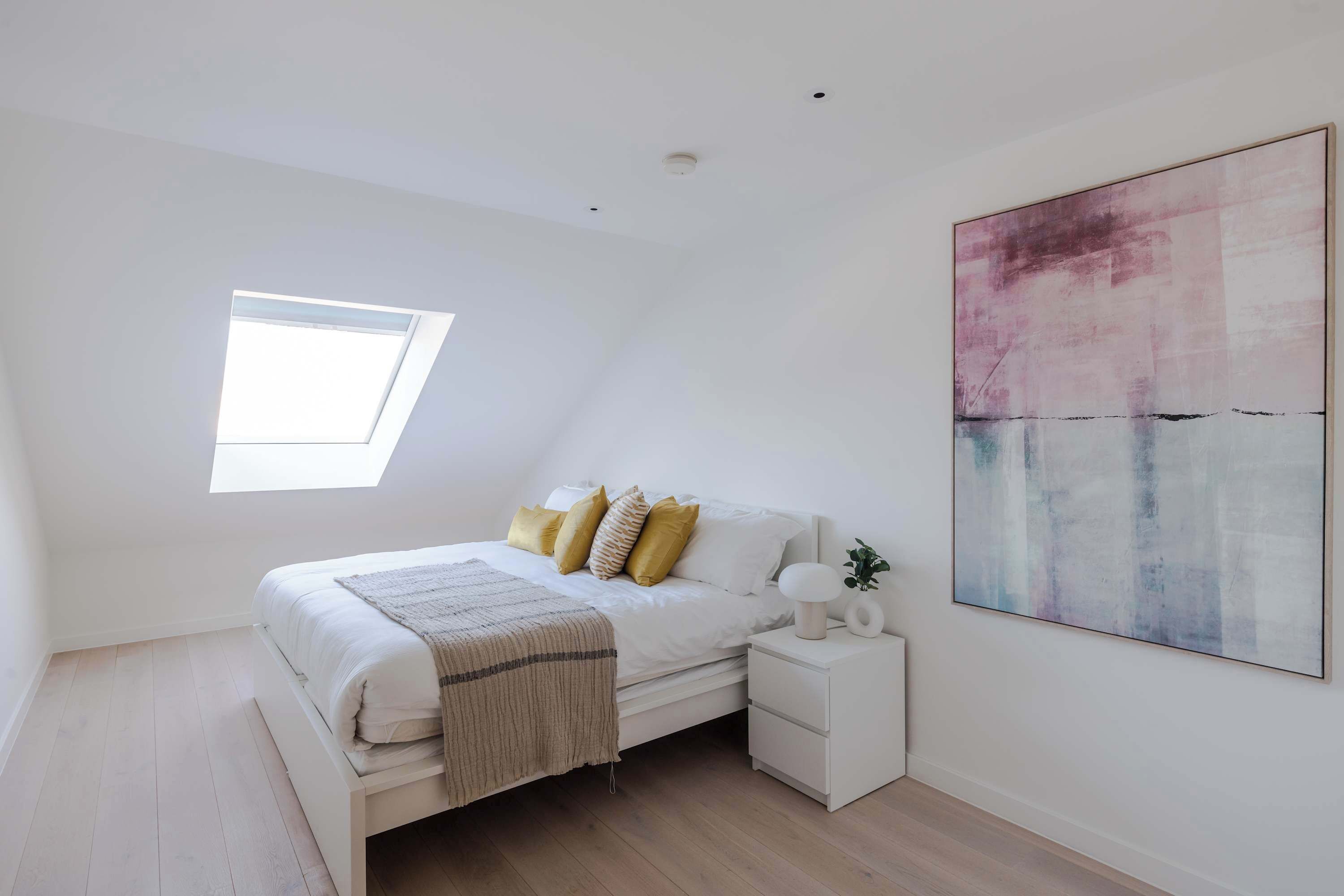 A bedroom with a skylight, a bed with white bedding and yellow pillows, a bedside table with a lamp, and a large abstract painting on the wall.