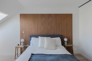 A bedroom with a wooden headboard, a bed with white bedding, two bedside tables with lamps, and a skylight.
