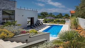 Un área de piscina moderna con una piscina azul cristalina, tumbonas y una escalera que conduce a una entrada cubierta.