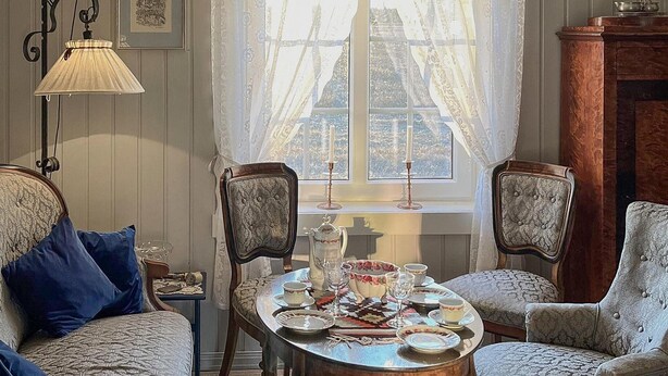 A cozy room with a dining table set for four, surrounded by patterned armchairs and a sofa with blue cushions. A window with sheer curtains allows natural light to fill the space. - 1 bedroom cozy home in Vormsund (Vormsund)