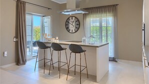Une cuisine moderne dotée d’un îlot central, de trois tabourets de bar, d’une horloge murale et d’une fenêtre avec des rideaux.