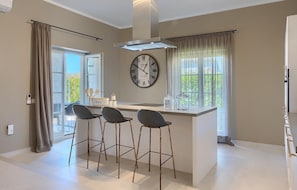 A modern kitchen with a central island, three bar stools, a wall-mounted clock, and a window with curtains.