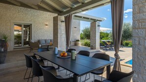 Une terrasse extérieure avec un mur en pierre, un espace repas couvert avec une table et des chaises, et une vue sur une piscine.