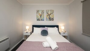 A neatly made bed with a pink blanket and a rolled towel, two bedside lamps, and two wall-mounted paintings of deer. - Yurali apartment is located within walking distance to the town centre. (Jindabyne)