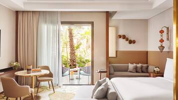 A hotel room with a bed, a sofa, a dining table with chairs, a small side table, and a view of a tropical garden through a glass door.