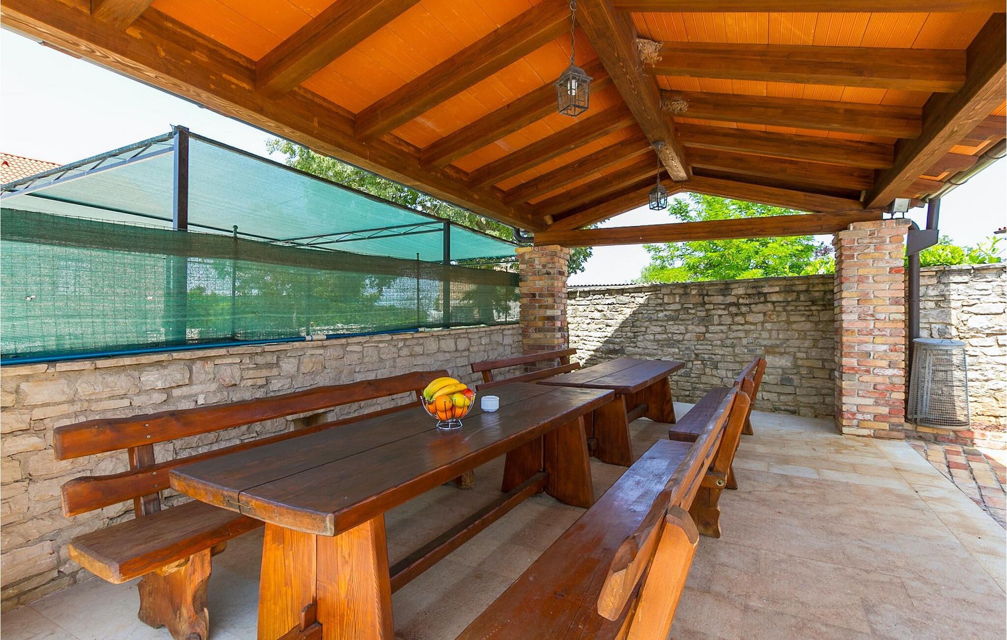 An outdoor patio with wooden benches and a table, covered by a wooden roof with a hanging lantern.