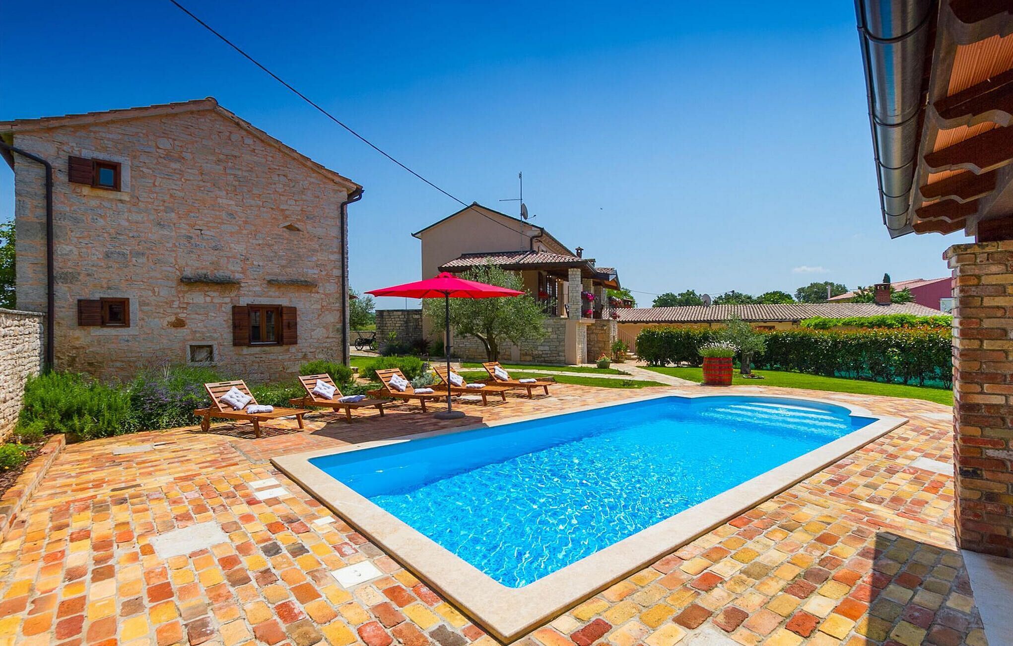 A swimming pool with lounge chairs and a red umbrella, surrounded by stone buildings and a well-maintained garden.