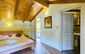 Habitación de hotel con techo de madera, cama con ropa blanca, mesita de noche, equipo de aire acondicionado y baño visible a través de una puerta abierta.