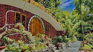 A brick building with a wooden door and windows, surrounded by lush greenery and a stone pathway.