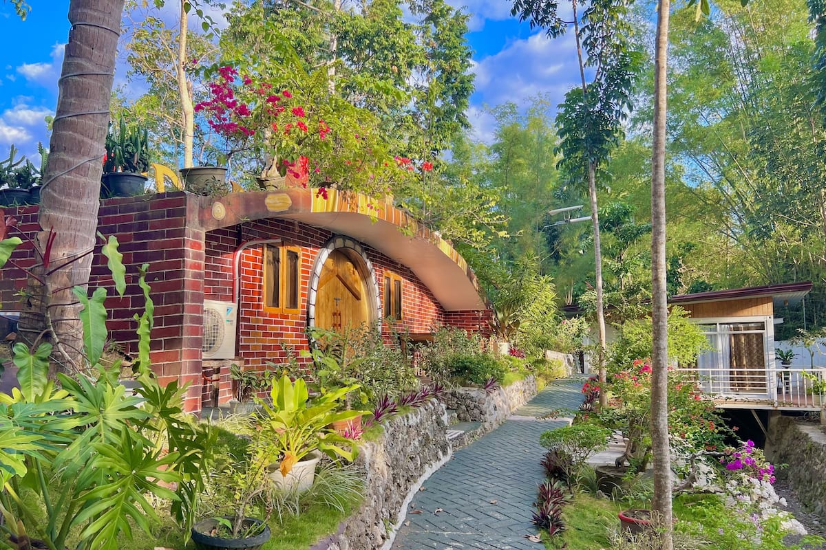A brick building with a curved archway, surrounded by lush greenery and potted plants.