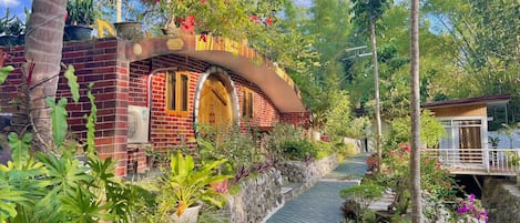 A brick building with a curved archway, surrounded by lush greenery and potted plants.