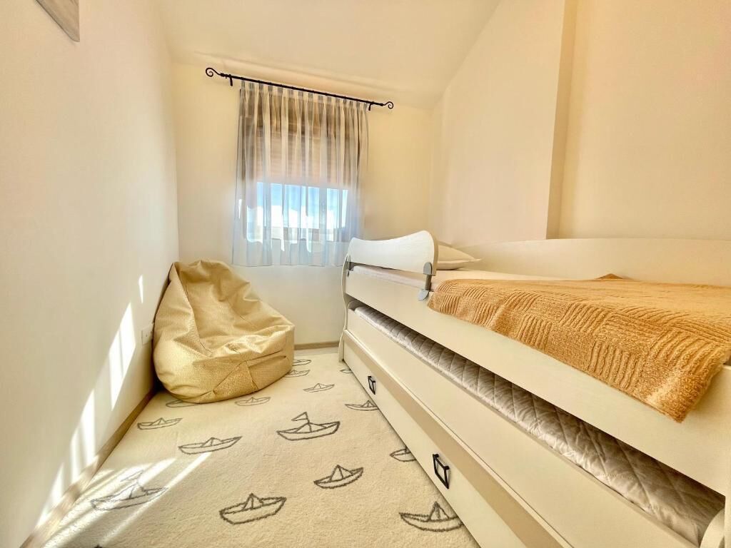 A small, cozy room with a bed, a window with curtains, and a patterned rug.