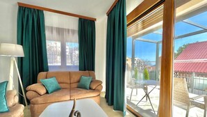 Ein modernes Wohnzimmer mit einem braunen Ledersofa, türkisblauen Kissen, einem weißen Couchtisch und einem großen Fenster mit grünen Vorhängen.