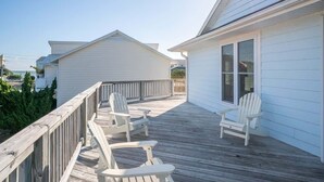 A wooden deck with white Adirondack chairs, a railing, and a building with a white exterior and large windows. - Sea La Vie Cottage Retreat (Kure Beach)