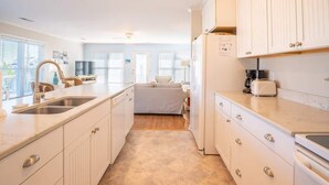 A modern kitchen with white cabinetry, a central island, and a dining area with a sofa. - Sea La Vie Cottage Retreat (Kure Beach)