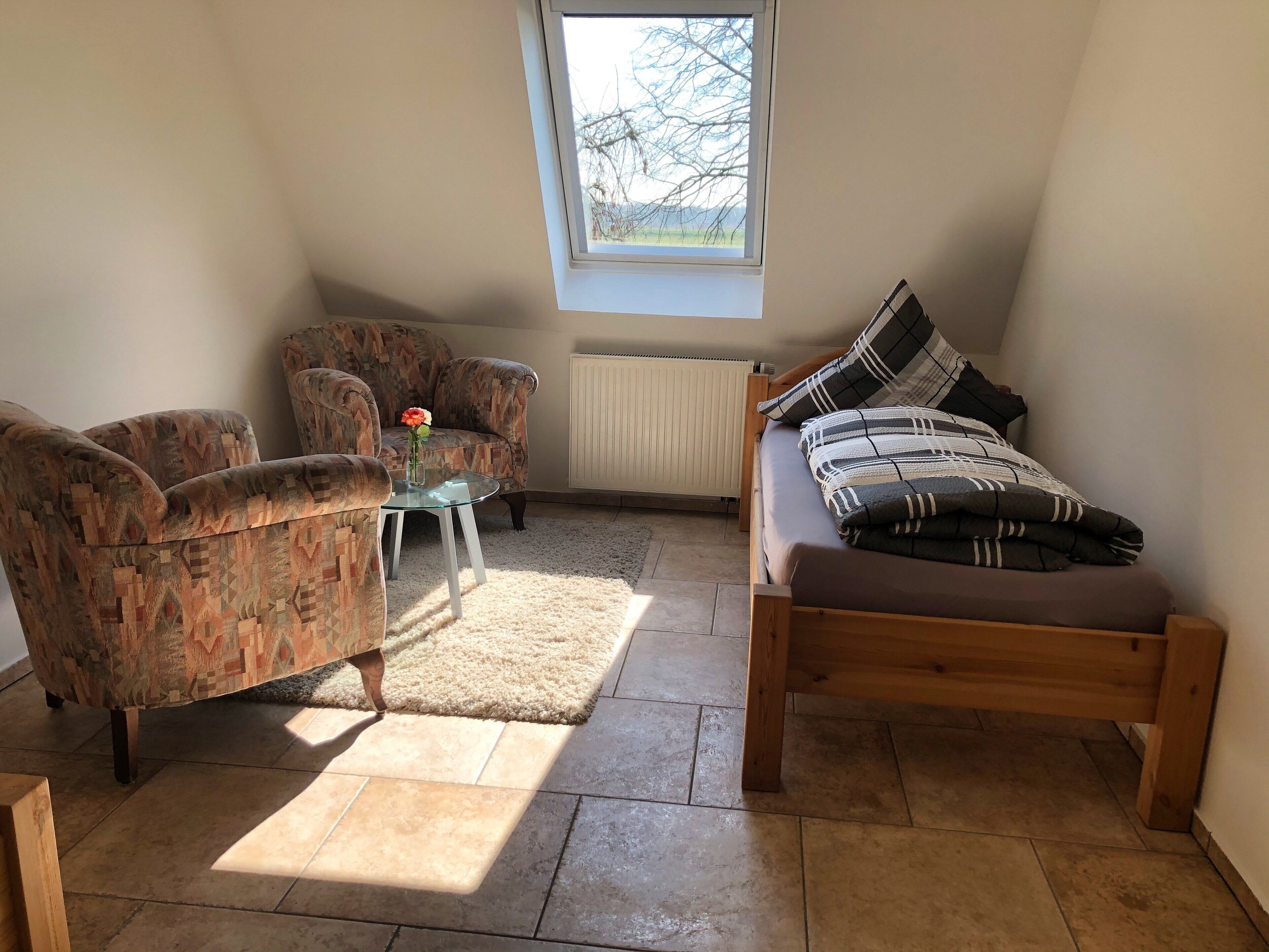 Un cuarto acogedor con cama, dos sillones, una mesita con un florero y vistas a árboles a través de un tragaluz.