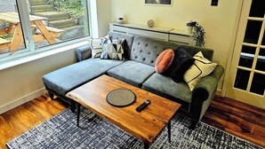Ein modernes Wohnzimmer mit einem grauen Ecksofa, einem Holztisch und einem großen Fenster mit Blick auf eine Terrasse.