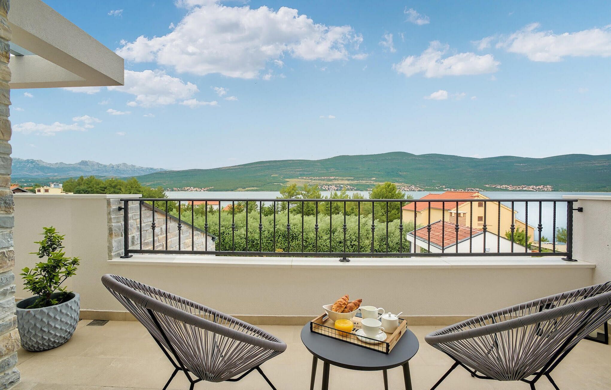 Ein Balkon mit Blick auf einen See und Berge, zwei Stühle, ein Tisch mit Frühstücksangeboten und ein Blumentopf.