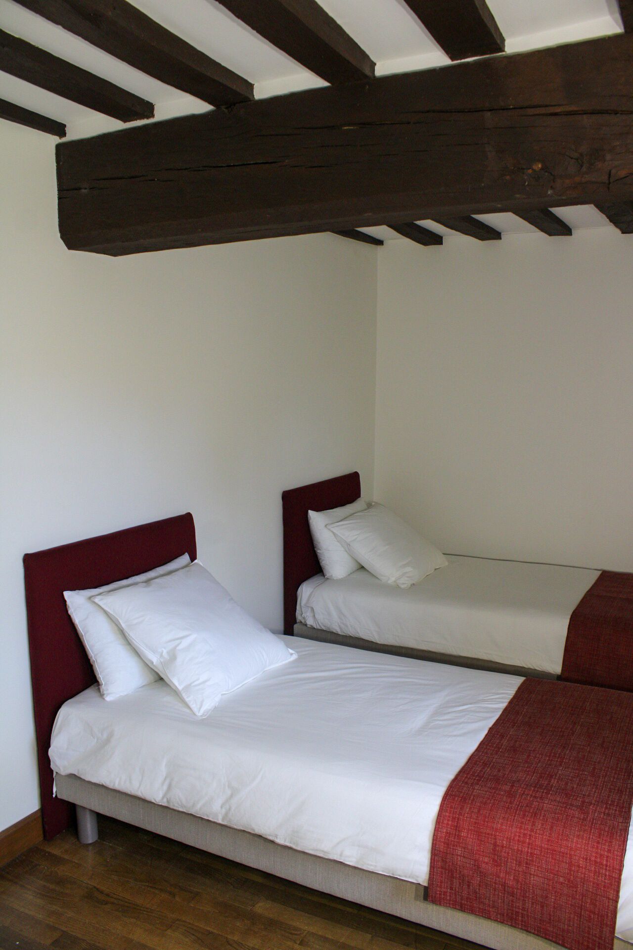 Deux lits simples avec du linge de lit blanc et des têtes de lit rouges, dans une pièce dont le plafond est orné de poutres en bois.