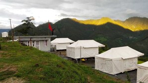 A row of white tents with a small building and a flagpole in the background, set against a mountainous landscape. - Khaltaal Camp And Resorts (Chamoli)