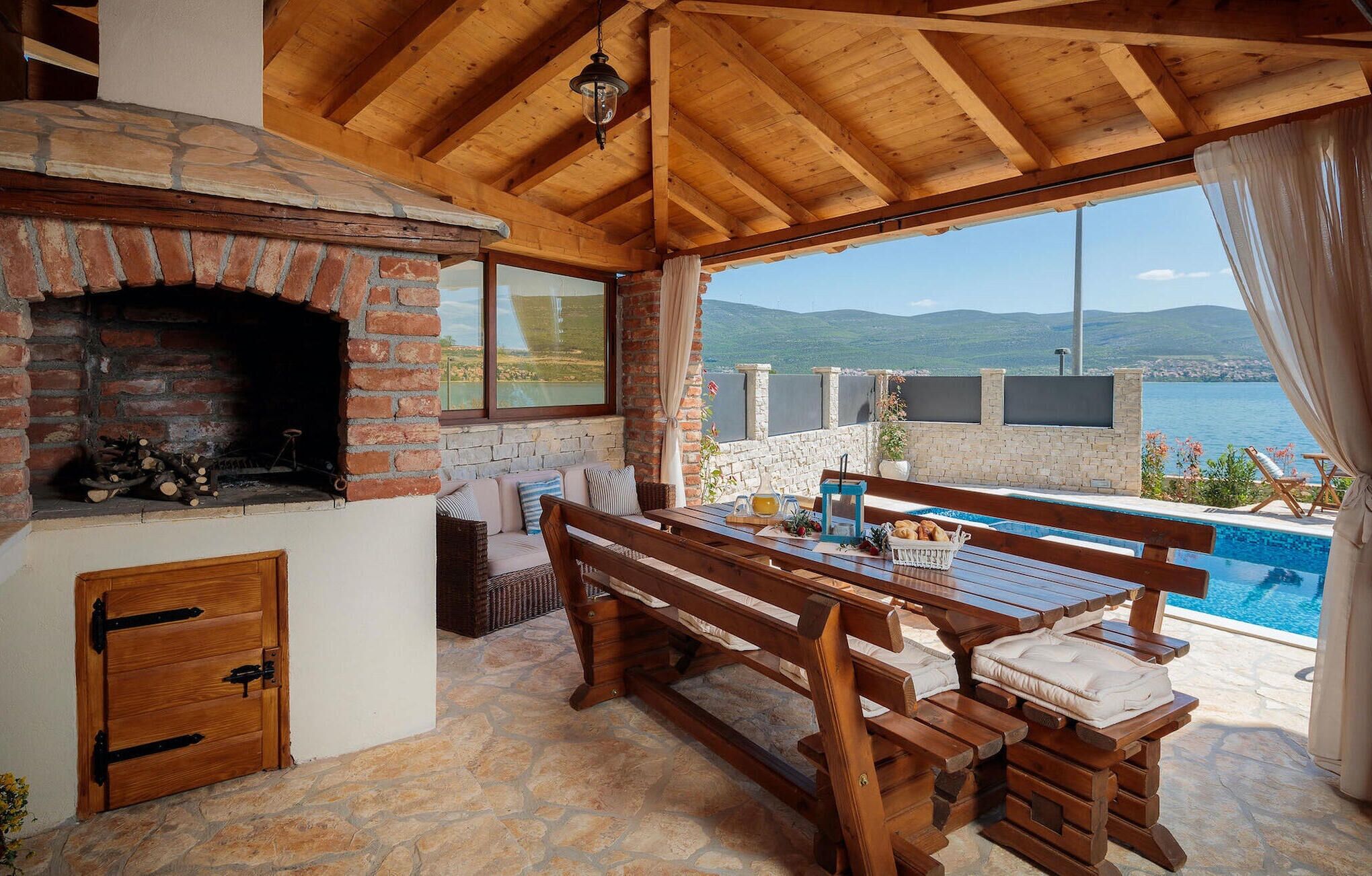 Un espace extérieur accueillant avec un coin salon, une cheminée en pierre, une piscine et une vue panoramique sur la mer et les montagnes.