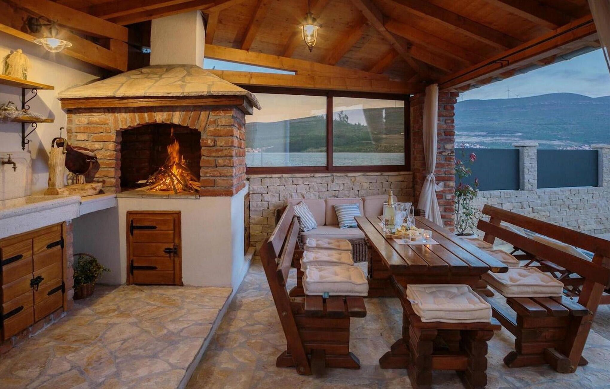 A rustic stone fireplace with a fire, wooden benches, a table with glasses, and a view of the sea and wind turbines.