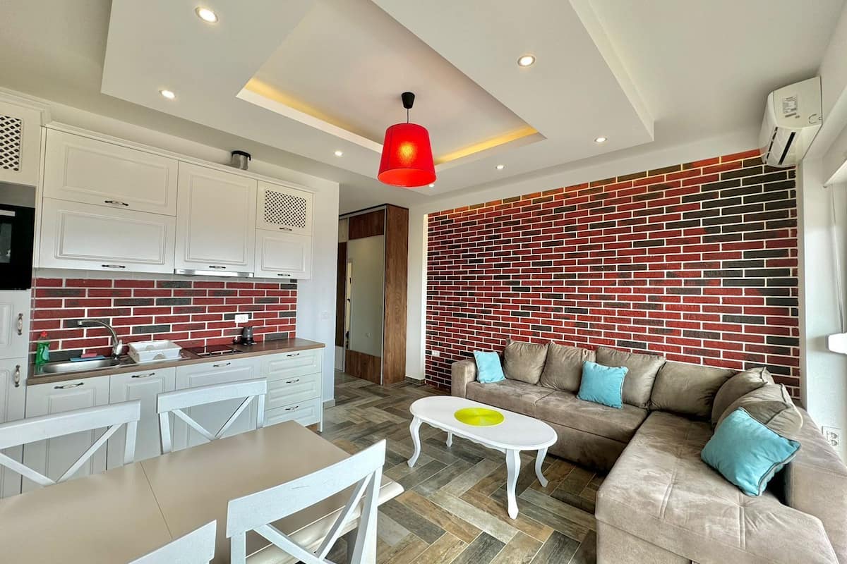 Una sala de estar moderna con sofá, una mesa de centro y una cocina con armarios blancos y un salpicadero de ladrillo rojo.